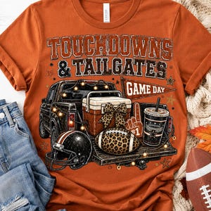 Puede incluir: Una camiseta color naranja quemado con un estampado gráfico de la caja de una camioneta con una hielera, un lazo de leopardo, un balón de fútbol americano de leopardo, un dedo de espuma y un banderín que dice GAME DAY. El texto dice TOUCHDOWNS & TAILGATES.