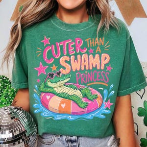 May include: Green t-shirt with the phrase "Cuter Than a Swamp Princess" in pink and orange. The graphic features a cartoon alligator wearing heart-shaped sunglasses and floating on a pink and orange inner tube in water.