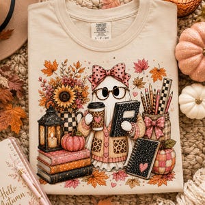 May include: A beige t-shirt featuring a graphic of a ghost wearing a leopard print cardigan, glasses, and a bow, holding a coffee cup and a book. The design includes sunflowers, a lantern, stacked books, pencils, and pumpkins surrounded by autumn leaves.