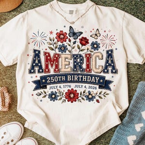 May include: White t-shirt with the word "AMERICA" in red, white, and blue lettering, celebrating the 250th birthday. The design includes flowers, butterflies, and fireworks. The dates "July 4, 1776" and "July 4, 2026" are also displayed.