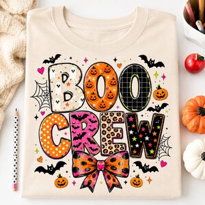 a t-shirt with a Halloween-themed design, including the word "BOO CREW" in a playful font, surrounded by various Halloween-related elements such as bats, pumpkins, and a bow.