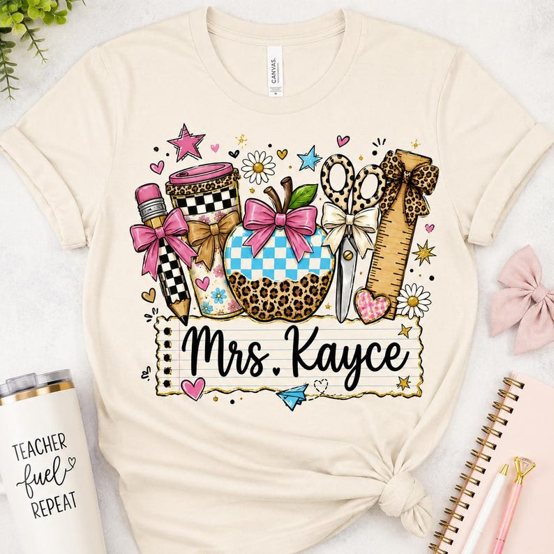 a white t-shirt with a colorful graphic design, including various school supplies and decorative elements such as a star, heart, and bow.