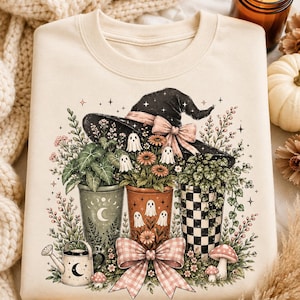 May include: A cream-colored crewneck sweatshirt featuring a graphic print of three potted plants, a large black witch hat with a pink bow, small white ghosts, a watering can, and a large pink gingham bow at the bottom with floral accents.
