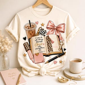 May include: A cream-colored short-sleeve t-shirt featuring a graphic print of an open book with the text I can do all things through Christ who strengthens me. Philippians 4:13, a coffee cup, a pink bow, a pen, a highlighter, and small hearts.
