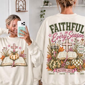 May include: A cream-colored crewneck sweatshirt featuring a graphic print of an open book, a cross, pumpkins, and autumn flowers. The back design includes the text FAITHFUL IN Every Season, GRACE UPON GRACE, and AUTUMN 2026 with the words GRACE, FAITH, HOPE, LOVE.
