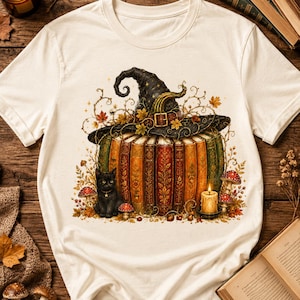 May include: A cream-colored short-sleeve t-shirt featuring a graphic print of a pumpkin constructed from vertical vintage books, topped with a black witch hat, surrounded by a black cat, autumn leaves, mushrooms, and a lit candle.