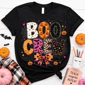 a black t-shirt with a Halloween-themed design, including the word "BOOCCREW" in a playful font, surrounded by various Halloween-related elements such as bats, pumpkins, and a spider web. The t-shirt is placed on a white surface, with additional Halloween decorations like a mug and a notebook nearby.