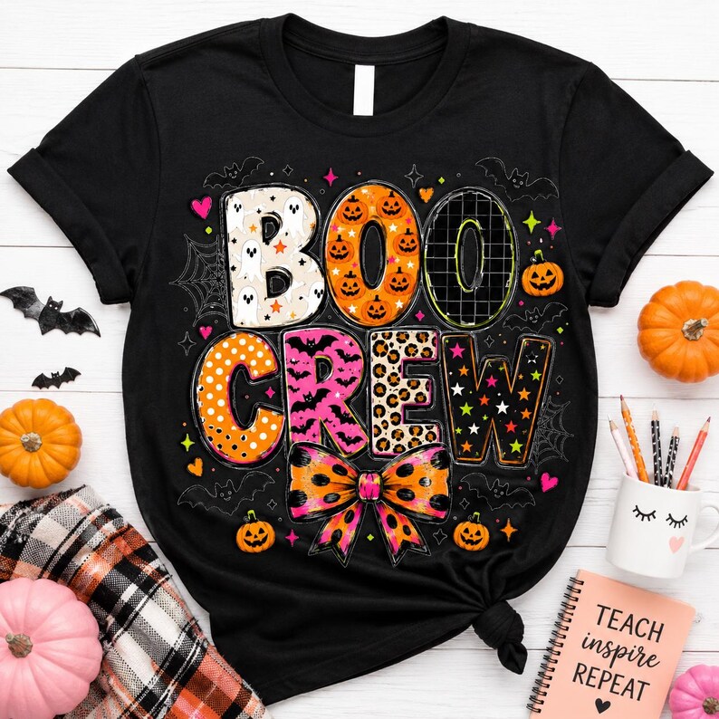 a black t-shirt with a Halloween-themed design, including the word "BOOCCREW" in a playful font, surrounded by various Halloween-related elements such as bats, pumpkins, and a spider web. The t-shirt is placed on a white surface, with additional Halloween decorations like a mug and a notebook nearby.
