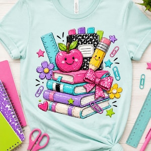 A light blue t-shirt with a colorful illustration of school supplies, including a smiling apple, books, and other school-related items, surrounded by various school supplies such as scissors, pens, and a ruler.
