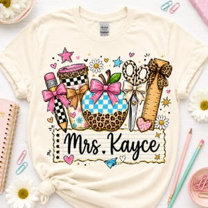 A white t-shirt with a colorful graphic design featuring various school supplies and the name "Mrs. Kayce" written on it.