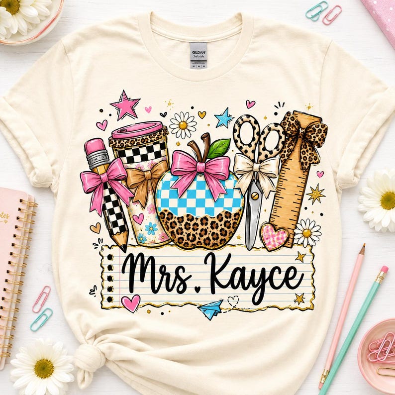 A white t-shirt with a colorful graphic design featuring various school supplies and the name "Mrs. Kayce" written on it.