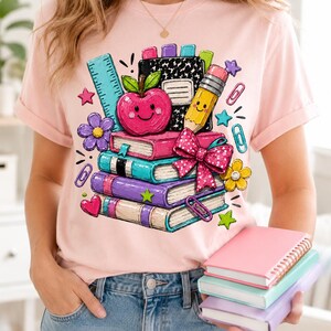 A woman wearing a pink t-shirt with a colorful graphic design of school supplies and books, holding a stack of books.