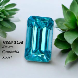 May include: A neon blue zircon gemstone with an emerald cut. The gemstone is a vibrant turquoise color and is set against a white background with green foliage. The text on the image reads "NEON BLUE Zircon Cambodia 3.55ct."