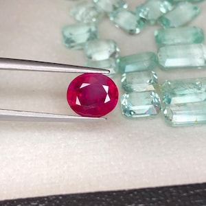 May include: A vibrant, round-cut ruby is held by tweezers, showcasing its deep red color and facets. Scattered around are various light blue-green gemstones, including rectangular and oval shapes, all on a neutral surface. The image highlights the beauty of the gemstones.