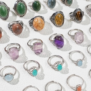 May include: A collection of silver-toned rings with various oval-shaped gemstones. The gemstones are in shades of green, purple, orange, yellow, and blue. The rings have ornate, detailed settings.
