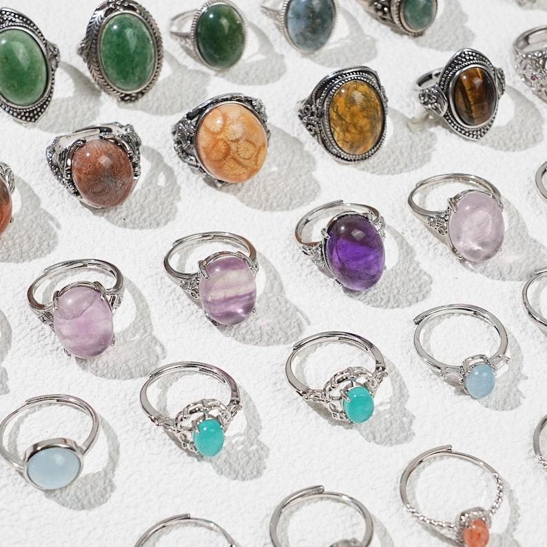 May include: A collection of silver-toned rings with various oval-shaped gemstones. The gemstones are in shades of green, purple, orange, yellow, and blue. The rings have ornate, detailed settings.