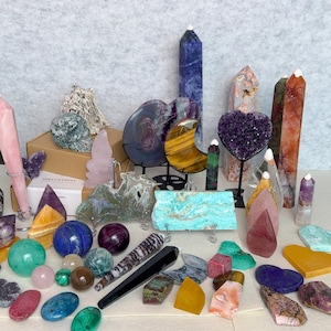May include: A collection of various polished and raw crystals, including towers, spheres, hearts, and slabs in colors like blue, purple, green, orange, and pink. The assortment features diverse mineral shapes, textures, and sizes arranged together.