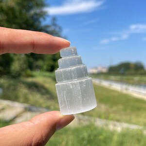 Può includere: Una torre di cristallo di selenite bianca traslucida intagliata in una forma cilindrica a gradini a più livelli. La pietra presenta una consistenza fibrosa e striata con una lucentezza naturale, tenuta tra due dita su uno sfondo esterno.