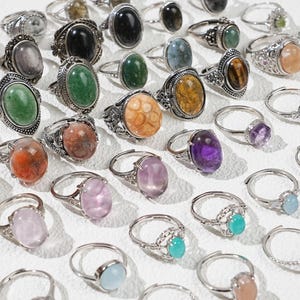 May include: A collection of gemstone rings with various oval and round stones in shades of green, purple, orange, and pink. The rings have silver-colored bands with ornate detailing. The gemstones are the focal point of the jewelry.