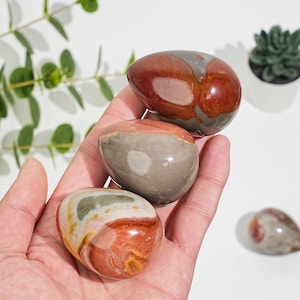 Può includere: Tre pietre lucide a forma di uovo, nei toni del rosso, grigio e crema, sono tenute in una mano. Le pietre hanno una finitura liscia e lucida e motivi naturali. Sullo sfondo c'è una piccola pianta succulenta in un vaso bianco.