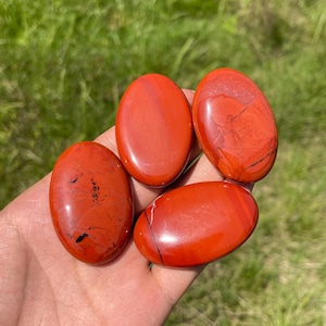 Può includere: Quattro pietre di diaspro rosso levigate e lisce di forma ovale, con variazioni naturali di colore che includono toni cremisi profondi, sottili riflessi arancioni e sottili venature nere sulla superficie delle pietre tenute nel palmo.