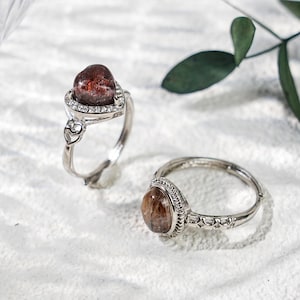 May include: Two silver rings with reddish-brown gemstones. One ring features a heart-shaped setting with a halo of small clear stones. The other ring has a round gemstone and a decorative band. The rings are displayed on a white surface.
