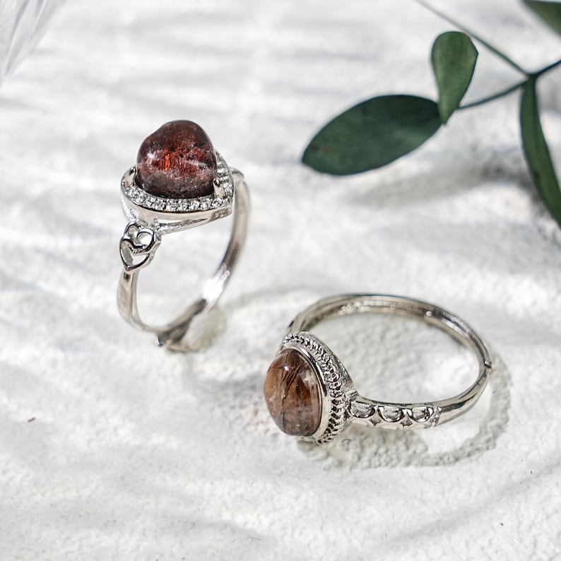 May include: Two silver rings with reddish-brown gemstones. One ring features a heart-shaped setting with a halo of small clear stones. The other ring has a round gemstone and a decorative band. The rings are displayed on a white surface.