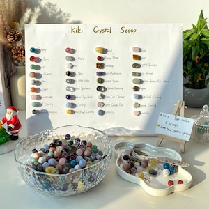 May include: A clear glass bowl filled with colorful crystal beads, alongside a white tray with a spoon and more crystals. A sign lists various crystal types, including spheres and worry stones. The sign reads "Kiki Crystal Scoop".