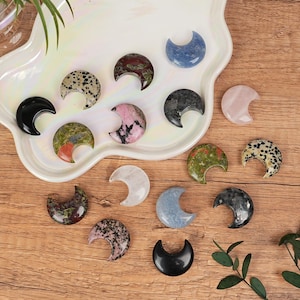 May include: A selection of crescent-shaped gemstones in diverse colours and patterns, such as black, pink, blue, and speckled. The gemstones are arranged on a white, scalloped tray and a wooden surface.