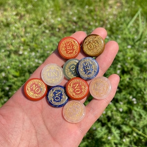 May include: A selection of small, circular stones, each displaying either "YES" or "NO" in gold script. The stones, in shades like red, blue, and gold, are held in a hand, with a blurred green background of grass.