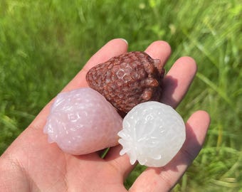 May include: Three small strawberry-shaped carvings made of polished stone. One is a deep reddish-brown, one is a pale translucent pink, and one is a clear, milky white. Each piece features a textured surface with small indentations and a leafy top.