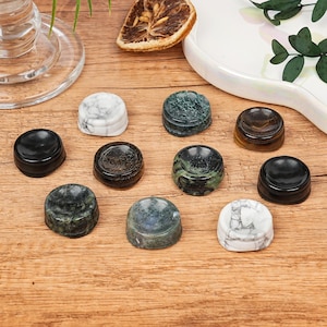 May include: A selection of small, round stone objects in various colours and patterns. The stones include black, white marble, and green speckled designs. The objects are arranged on a wooden surface, with decorative elements in the background.