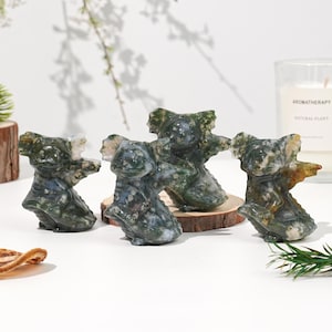 Puede incluir: Cuatro figuras de koalas de ágata musgosa. Los koalas están tallados en piedra verde, blanca y marrón y se exhiben sobre pequeños trozos de madera. El fondo incluye una vela y vegetación.