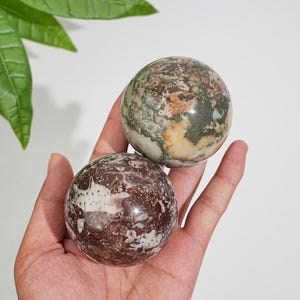 Puede incluir: Dos esferas de piedra pulida. La esfera superior presenta un patrón moteado en tonos verde oliva, crema y marrón rojizo. La esfera inferior muestra una base de color granate oscuro y marrón con intrincados patrones de vetas blancas y beige.