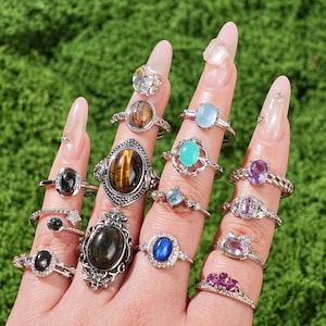 May include: A hand displays a collection of gemstone rings. The rings feature various stones, including black onyx, tiger's eye, and amethyst, set in silver bands. Some rings have intricate designs, while others are more minimalist. The background is a vibrant green.