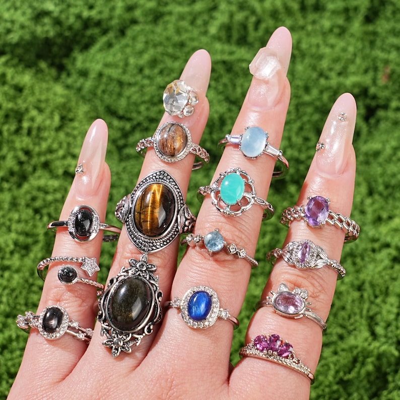May include: A hand displays a collection of gemstone rings. The rings feature various stones, including black onyx, tiger's eye, and amethyst, set in silver bands. Some rings have intricate designs, while others are more minimalist. The background is a vibrant green.