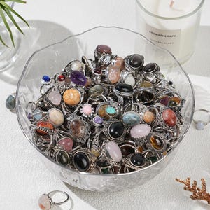 Puede incluir: Un cuenco de vidrio transparente lleno de numerosos anillos de piedras preciosas de varios colores. Los anillos tienen engastes de metal plateado y presentan piedras negras, rosas, marrones y azules. Una vela encendida con el texto "AROMATHERAPY NATURAL PLANT" es visible.