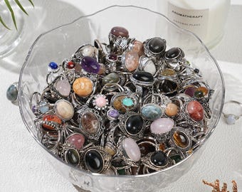 May include: A clear glass bowl overflowing with a collection of gemstone rings. The rings feature various stone colors, including black, pink, brown, and blue, set in silver-toned metal. A lit candle with the text "AROMATHERAPY NATURAL PLANT" is in the background.