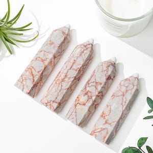 Puede incluir: Cuatro obeliscos con vetas de mármol con base blanca y punta. Los obeliscos tienen un fondo gris claro con vetas rojas. Los objetos están dispuestos en diagonal sobre una superficie blanca.