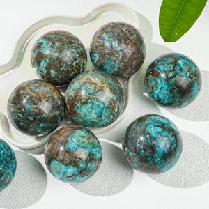 May include: A collection of polished gemstone spheres, each displaying a mix of teal, brown, and gray hues. The spheres are arranged on a white, scalloped dish, showcasing their natural patterns and textures. The overall aesthetic is earthy and elegant.