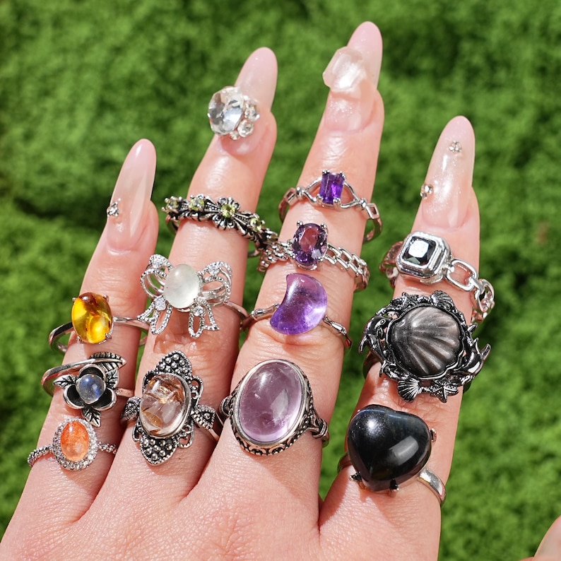 May include: A collection of gemstone rings in various designs and colors, including clear, purple, yellow, and black stones. The rings are displayed on a hand against a green background, showcasing a variety of styles and settings.