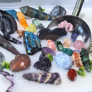 May include: A collection of polished gemstones and crystals in various shapes and colors, including amethyst, labradorite, and rose quartz. A small pink elephant figurine is also present. The stones are arranged on a bed of white beads.