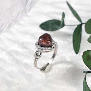 May include: A silver ring featuring a heart-shaped, dark red gemstone. The gemstone is surrounded by a halo of small, clear stones. The ring's band has a delicate design with heart-shaped accents, creating a romantic and elegant aesthetic.