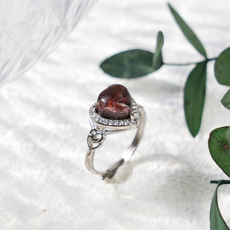 May include: A silver ring featuring a heart-shaped, dark red gemstone. The gemstone is surrounded by a halo of small, clear stones. The ring's band has a delicate design with heart-shaped accents, creating a romantic and elegant aesthetic.