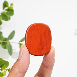Puede incluir: Una piedra de jaspe rojo de forma ovalada, pulida y lisa. La piedra presenta tonos rojo anaranjados profundos y sutiles vetas naturales oscuras y finas que recorren su superficie, mostrando un acabado brillante y una forma redondeada y plana.