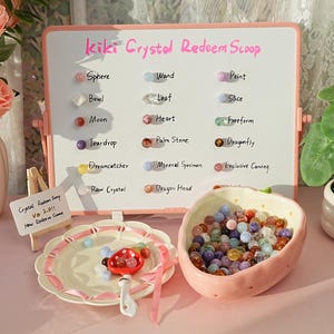 Puede incluir: Una presentación con una pizarra que muestra "kiki Crystal Redeem Scoop" y formas de cristal. Un cuenco con forma de fresa contiene cristales de colores, y también hay un plato con cristales y una cuchara.