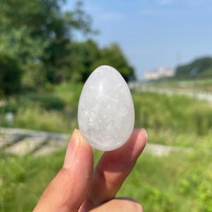 Puede incluir: Un cristal de cuarzo blanco pulido en forma de huevo con apariencia translúcida y fracturas internas, sostenido frente a un entorno natural al aire libre. La piedra presenta una parte superior redondeada y una base más ancha, con textura clara.