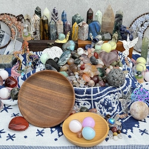 May include: A collection of various crystals and gemstones, including polished stones, crystal points, and spheres. The arrangement includes a wooden plate and a small wooden bowl with colorful spheres. The background features a patterned textile.