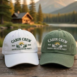 May include: Two baseball caps, one white and one olive green, with the text "CABIN CREW" above a mountain and sun graphic. The text "MAY 2026" and "DAVID'S BACHELOR PARTY" are also printed on the caps. The caps are on a wooden surface.