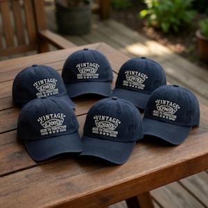 May include: A collection of navy blue baseball caps with white text. Each cap features the word "VINTAGE" above a year, such as 1985, 1992, 1978, 1969, 2001, and 2010, with the phrase "AGED TO PERFECTION" below.